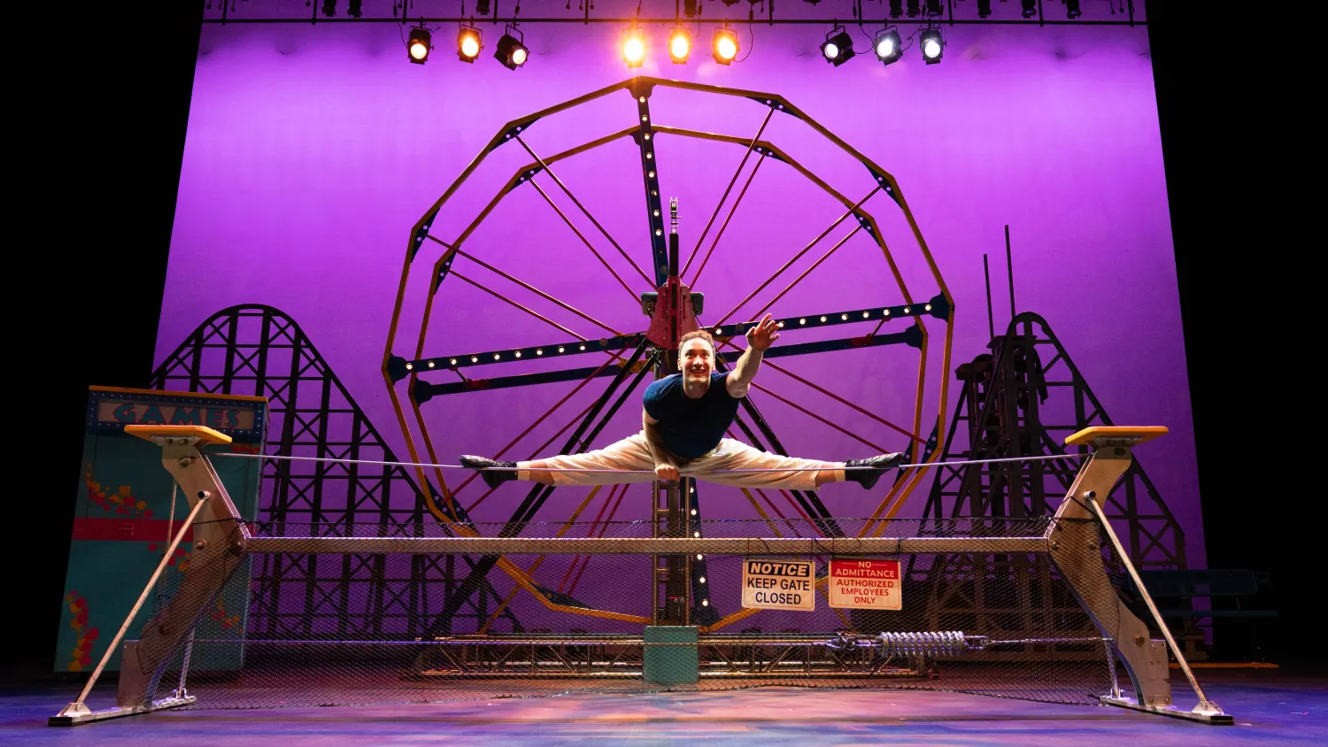 A man balances in a straddle on a tight wire and reaches one hand towards the audience, smiling.