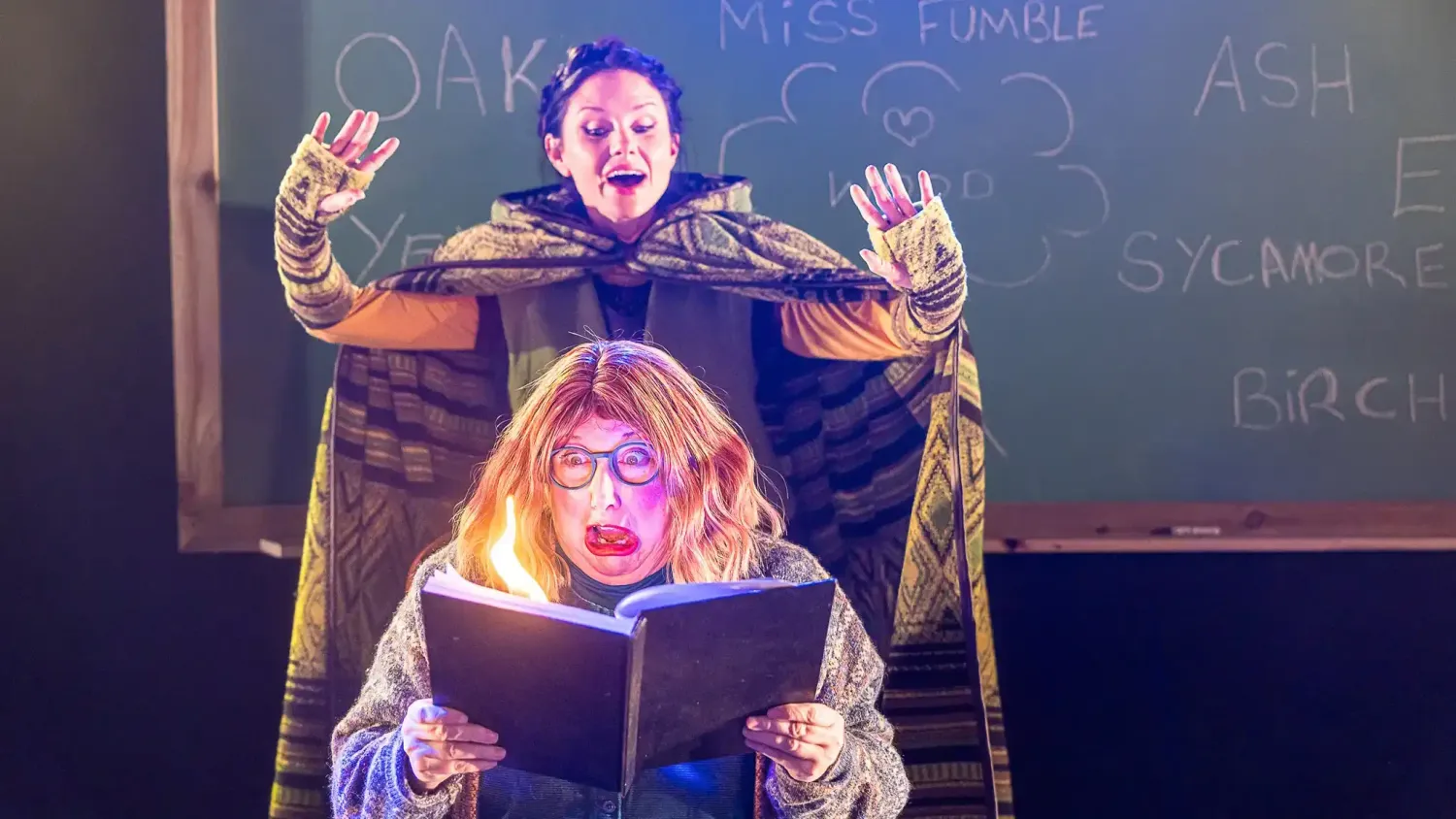 A performer wearing glasses holds a book and makes a shocked face as the performer behind them wearing a cloak sets fire to a page of the book.