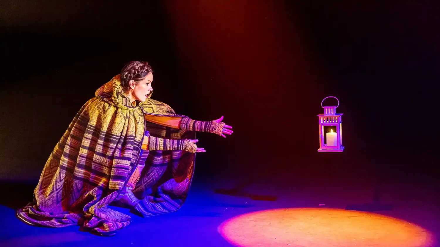 A performer wearing a rich patterned cloak crouches and reaches out towards a floating lantern.