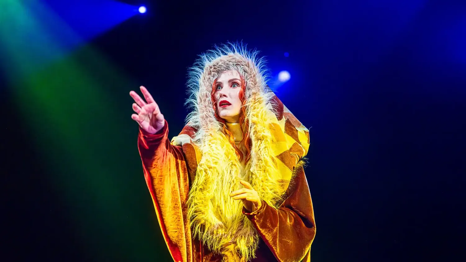 A performer with long red hair wears a patchwork hood trimmed with fur and reaches out with one hand.