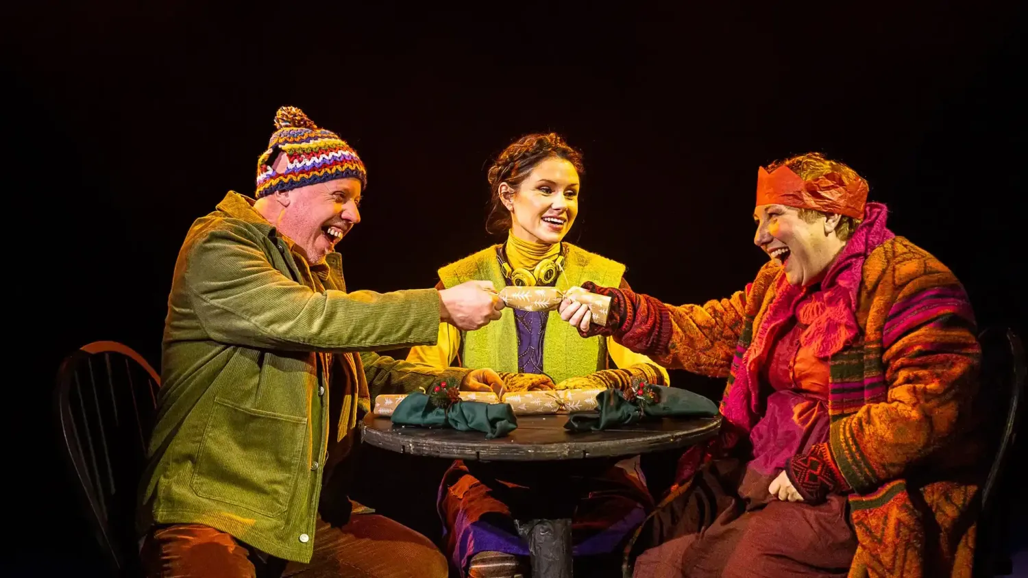 Three performers in warm clothing sit at a small round wooden table smiling as they open holiday crackers.