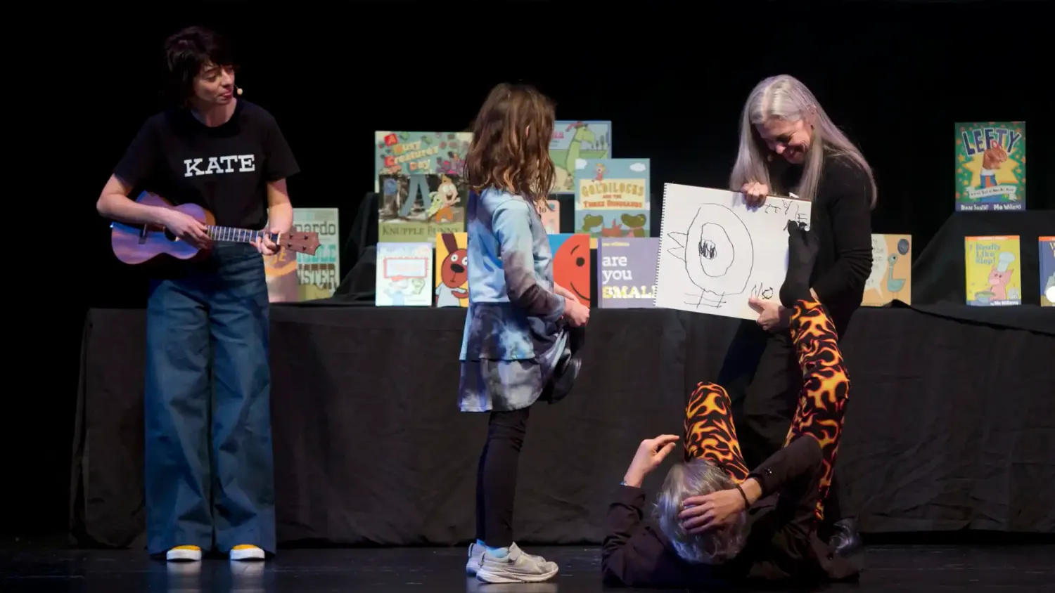 A performer stands on the left holding a ukulele. A child stands on stage watching a performer in fire pants draw a pigeon with their feet and a third performer holds the sketchpad.