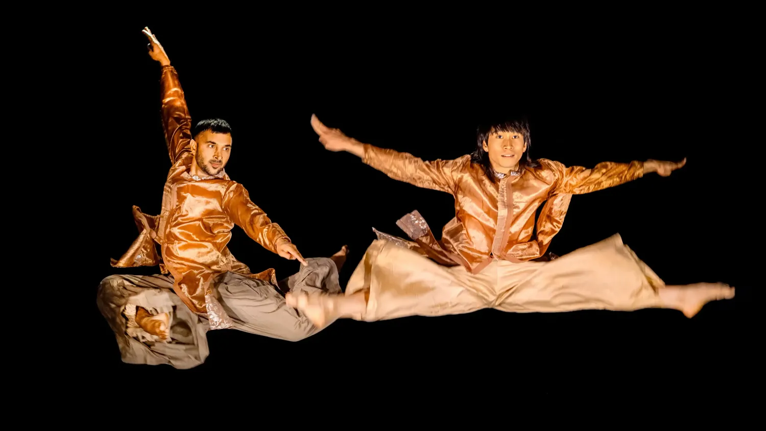 Two dancers in shiny brown clothing leap in the air with their arms and legs outstretched.