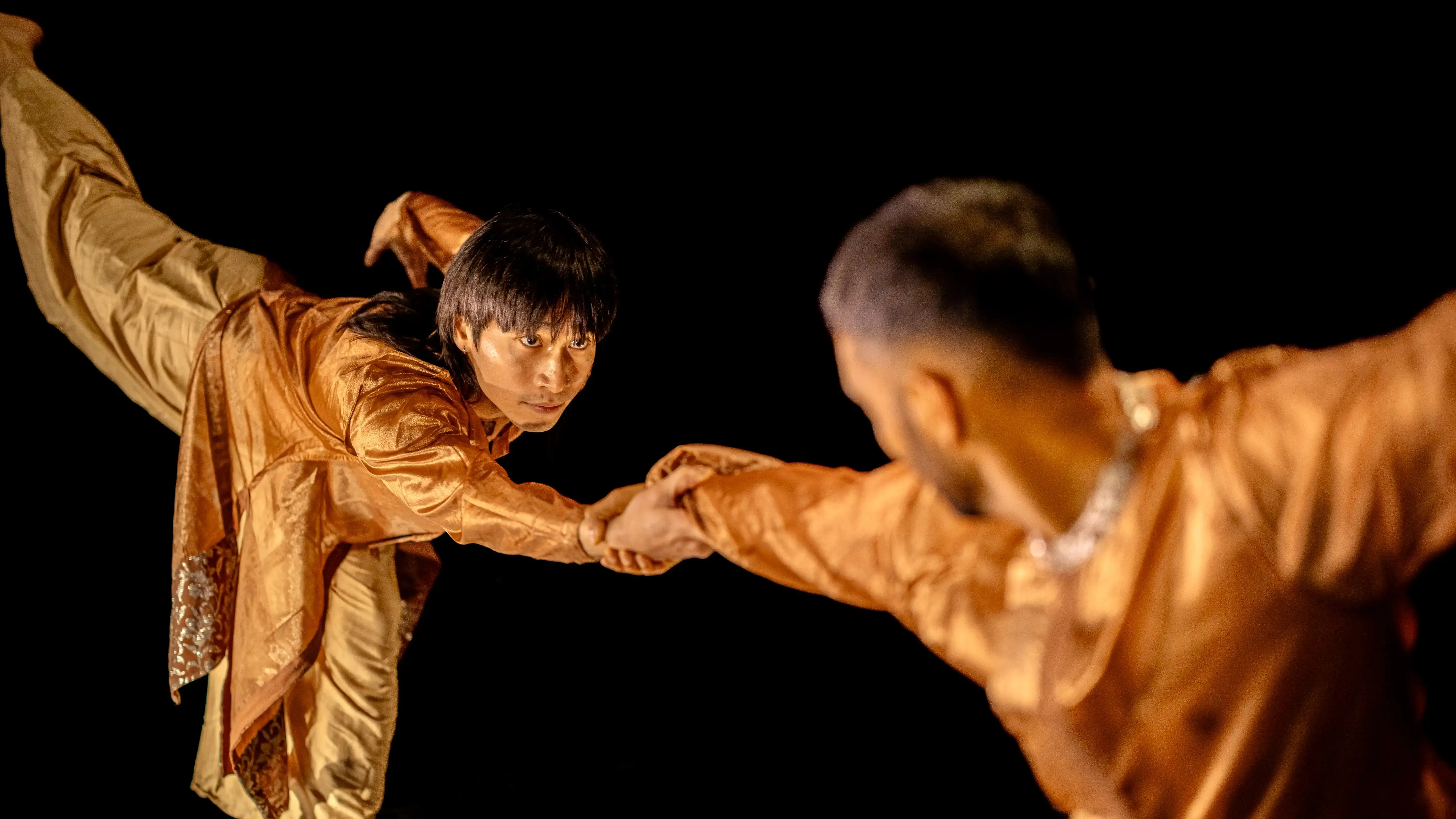 Two dancers reach out to grasp each other's hand and lean forward, raising their back foot towards the sky.