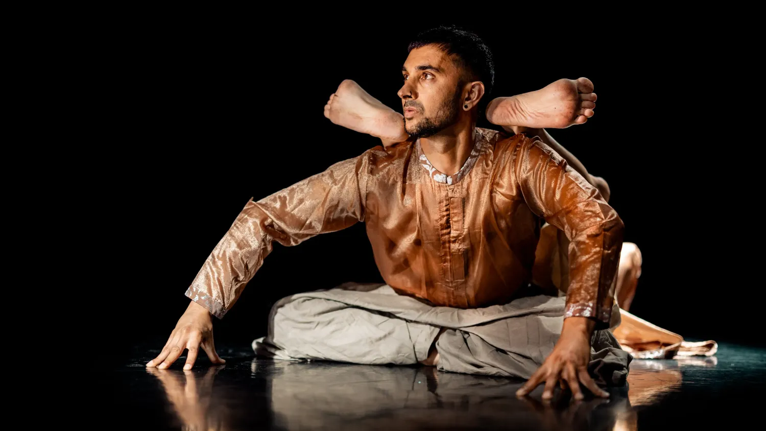 A dancer in shiny brown clothing looks left, sits and leans forward on his hands. Another dancer lays back and rests his feet on the first dancer's shoulders.
