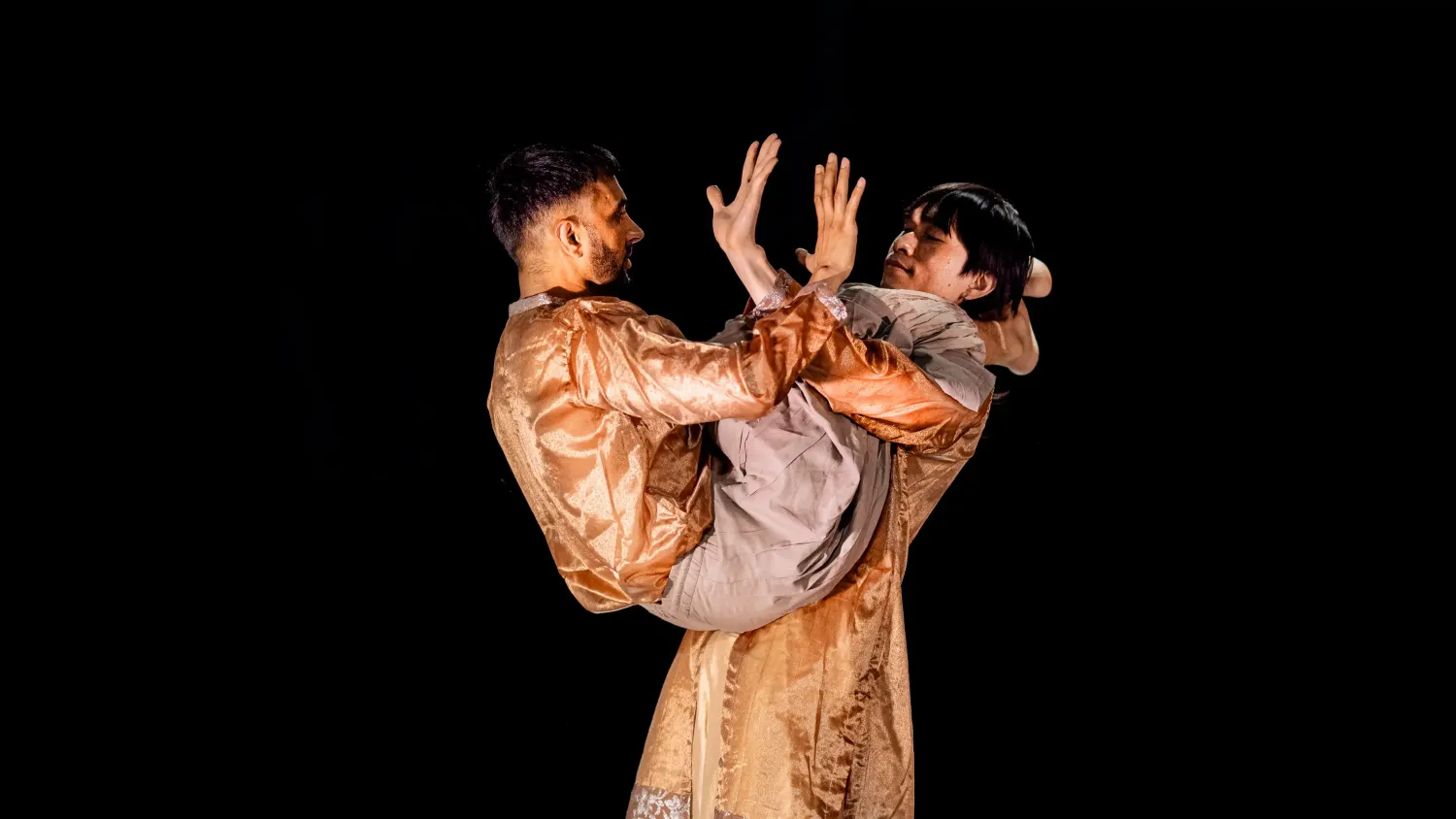 One dancer stands holding the other, who wraps his legs around the standing dancer's neck. They show each other the palms of their hand.