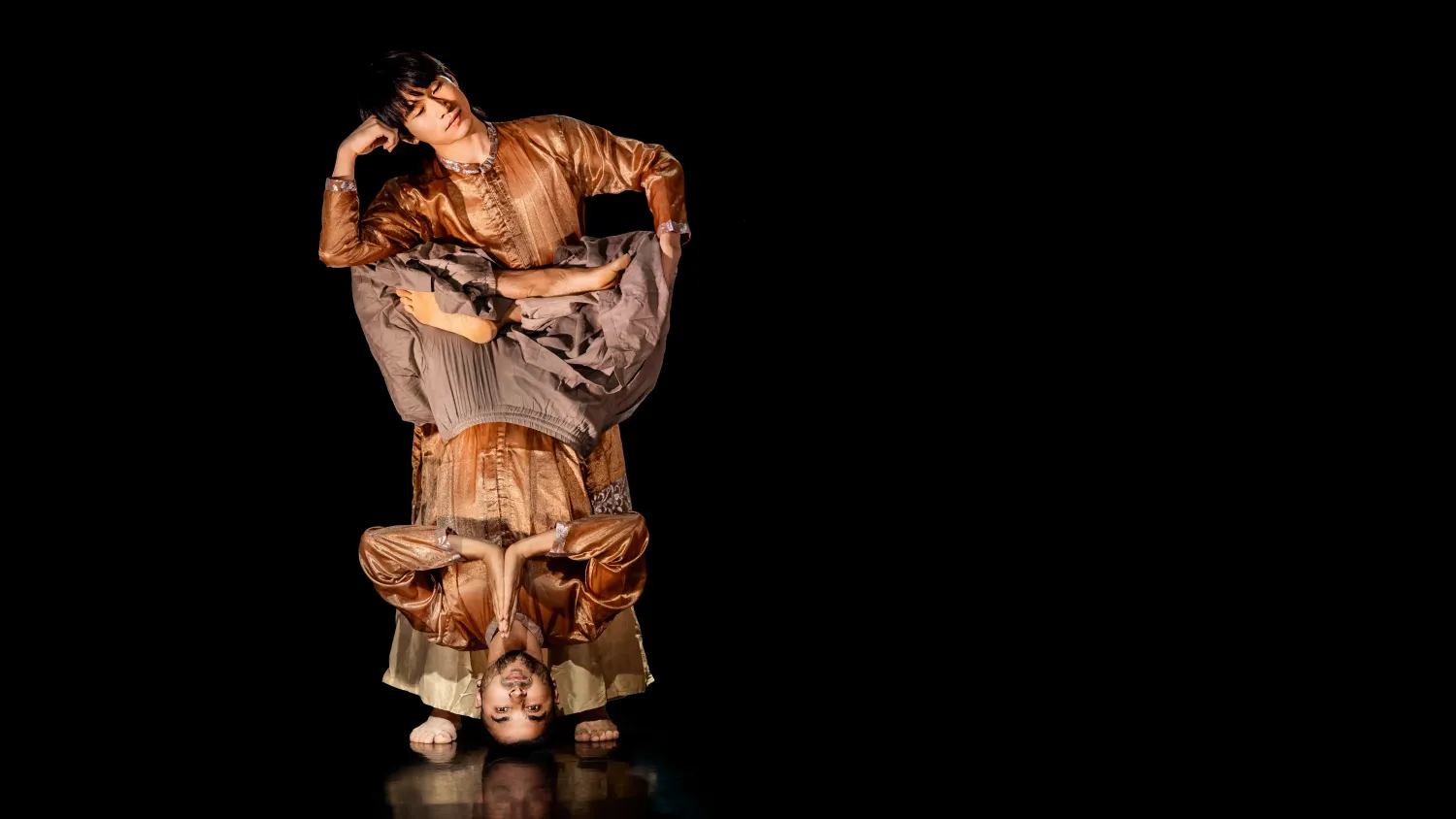 A dancer does a headstand with his legs folded and holds his palms together. A second dancer stand behind him and rests his head on his arm, which rests on the first dancer's legs.