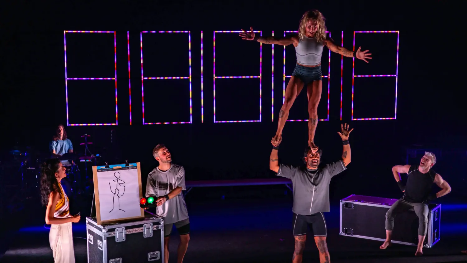 A woman stretches her arms out and balances standing on a man's head to match a stick figure drawing next to them. The man holds one hand freely above his head.