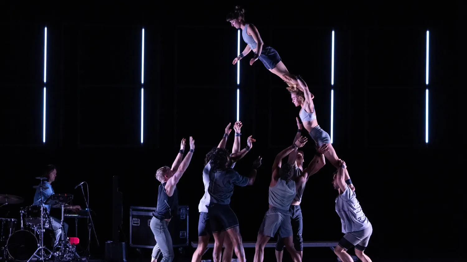 Three performers stacked on each other's shoulders lean toward the ground as five other performers prepare to catch them.