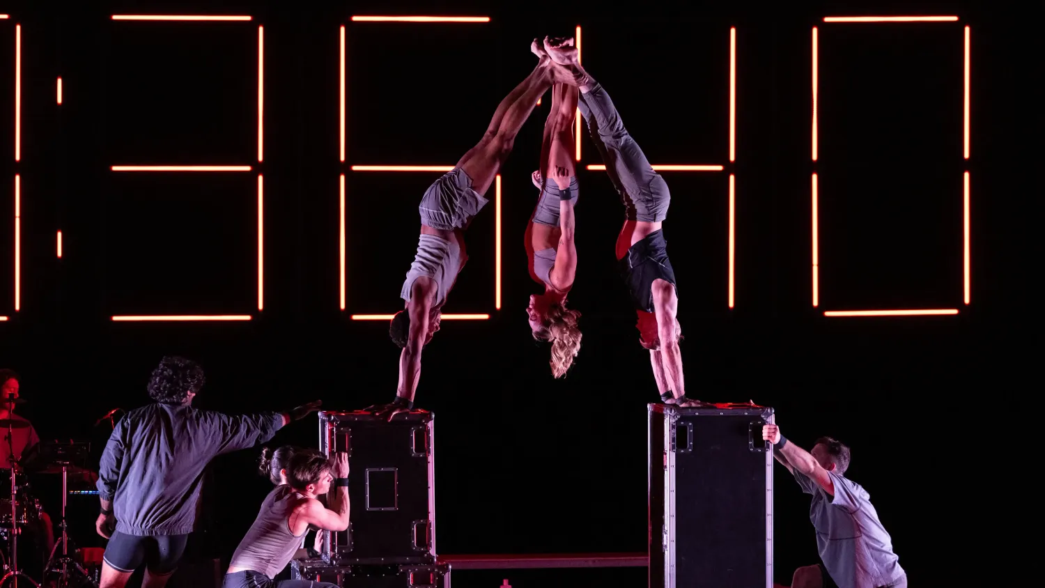 Two performers handstand on boxes, facing one another and holding their feet together. A third performer hangs upside down between them.