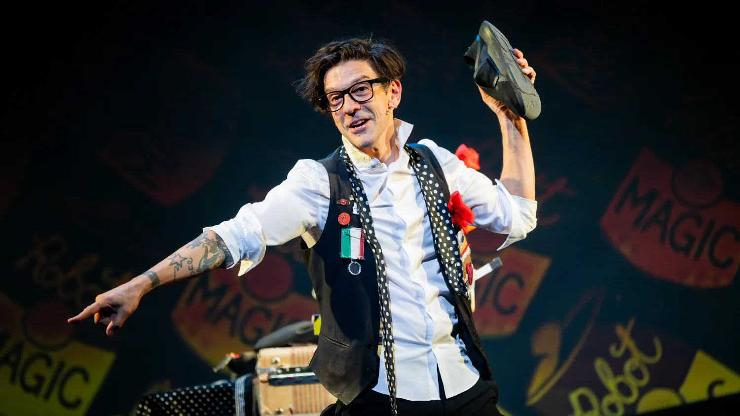 A magician holds a black sneaker in his right hand and points down with his left hand.