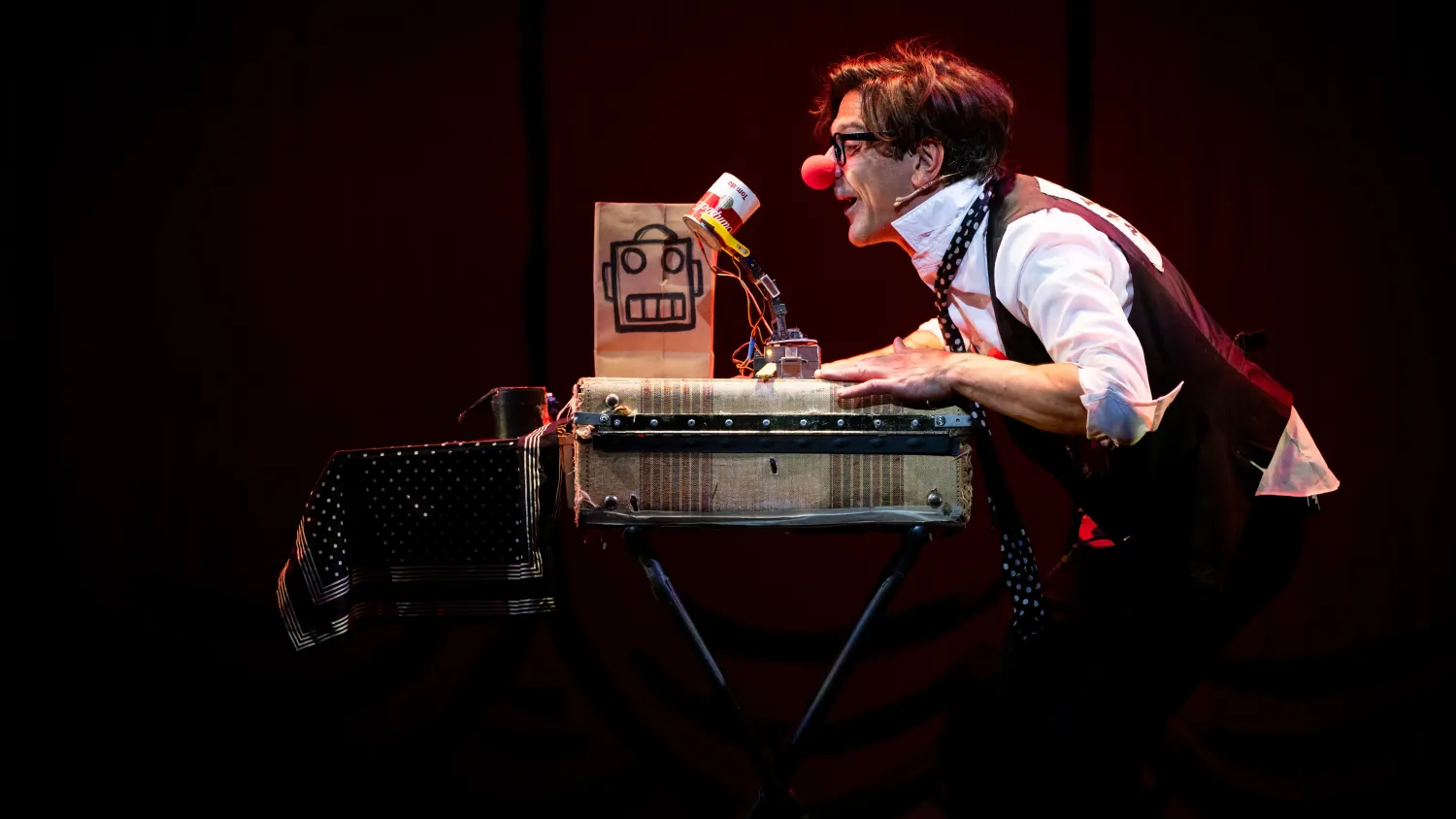 A man wearing a red clown nose leans forward on a briefcase towards a small robot made from a mechanical arm and tomato soup can.
