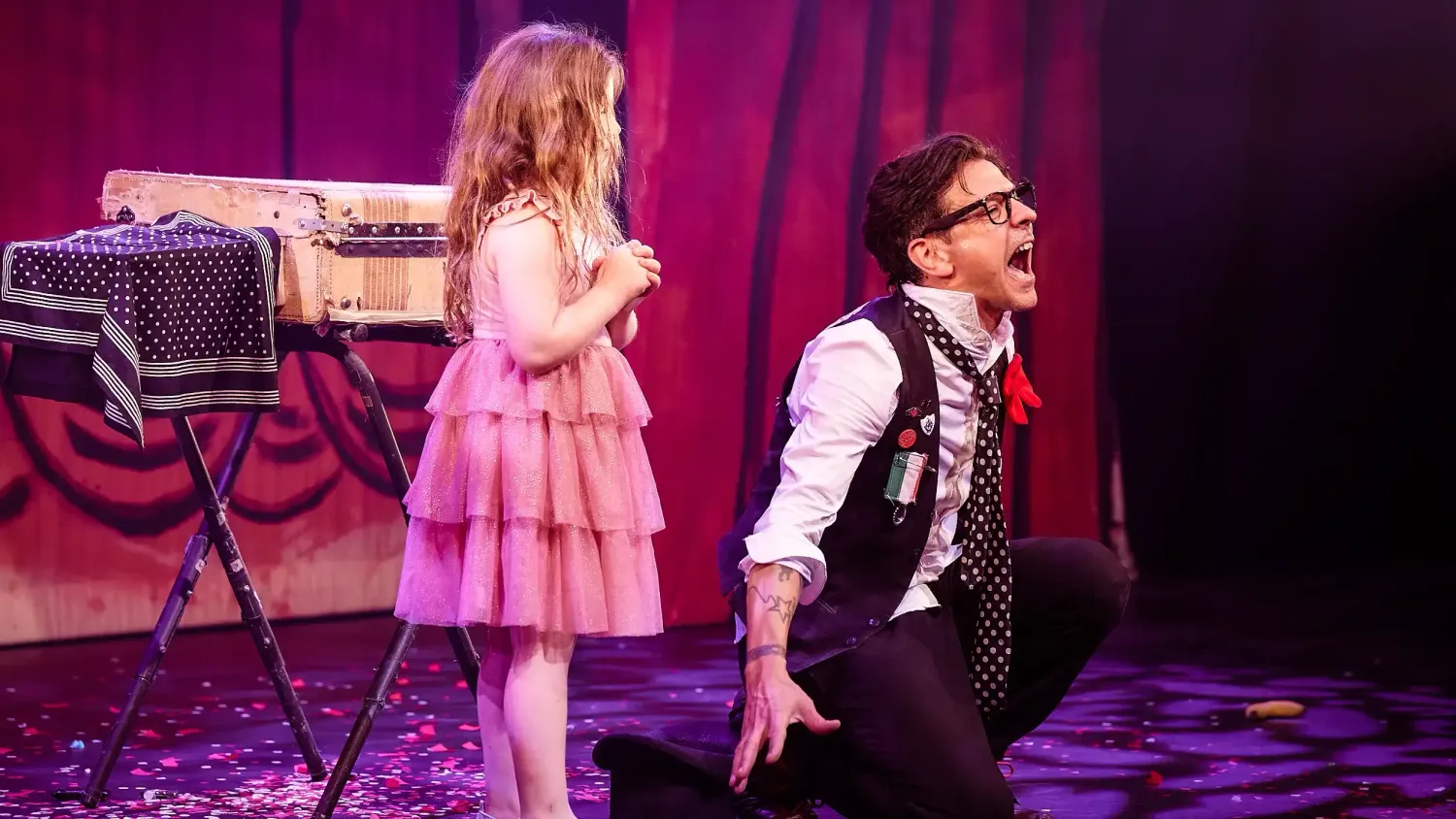 A young girl with long hair and a pink dress stands on stage. On her right, a man crouches and screams out towards the audience.