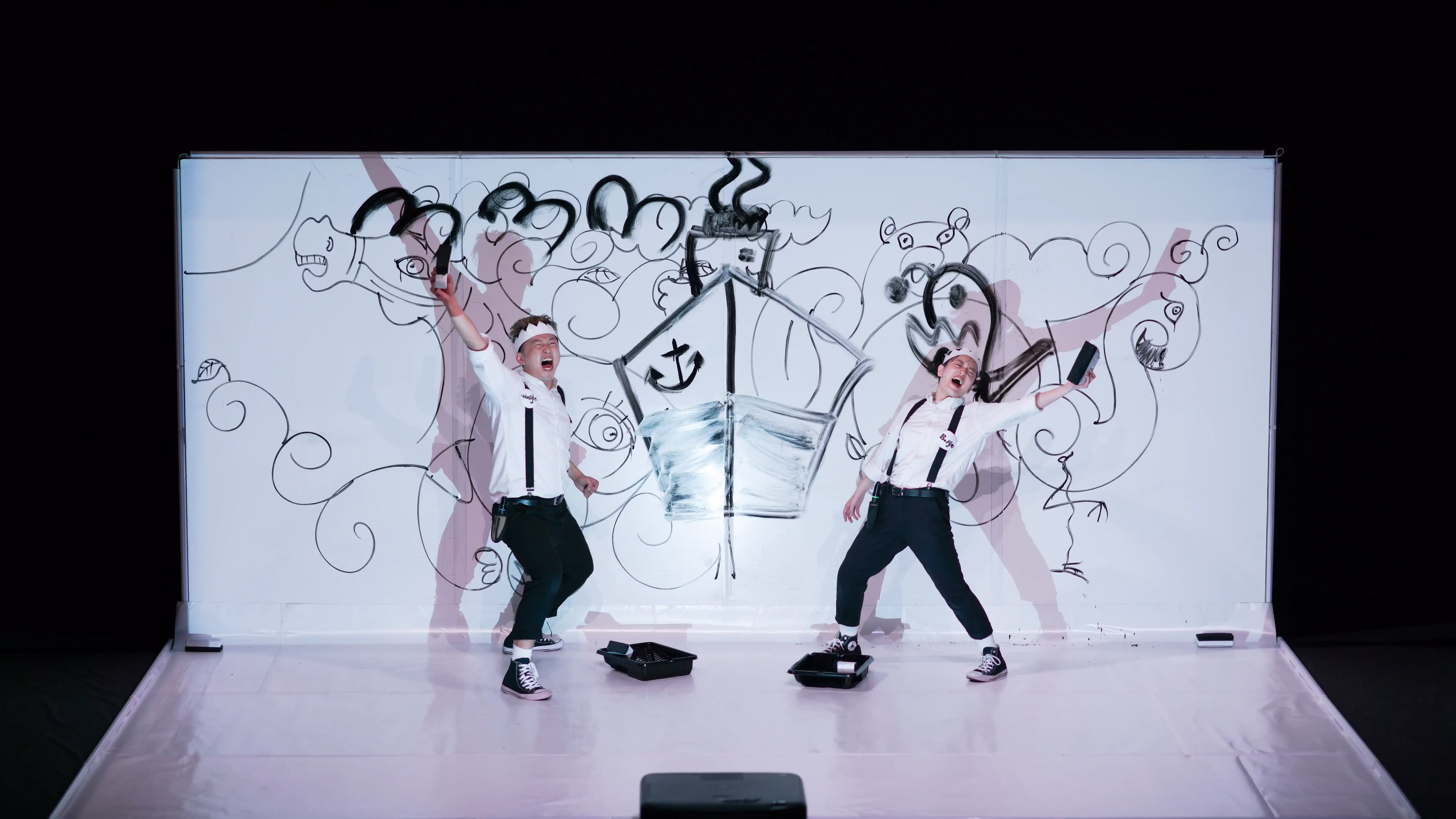 Two performers wearing suspenders and paper crowns exclaim and strike a pose with one arm raised in front of a large white dry erase board.
