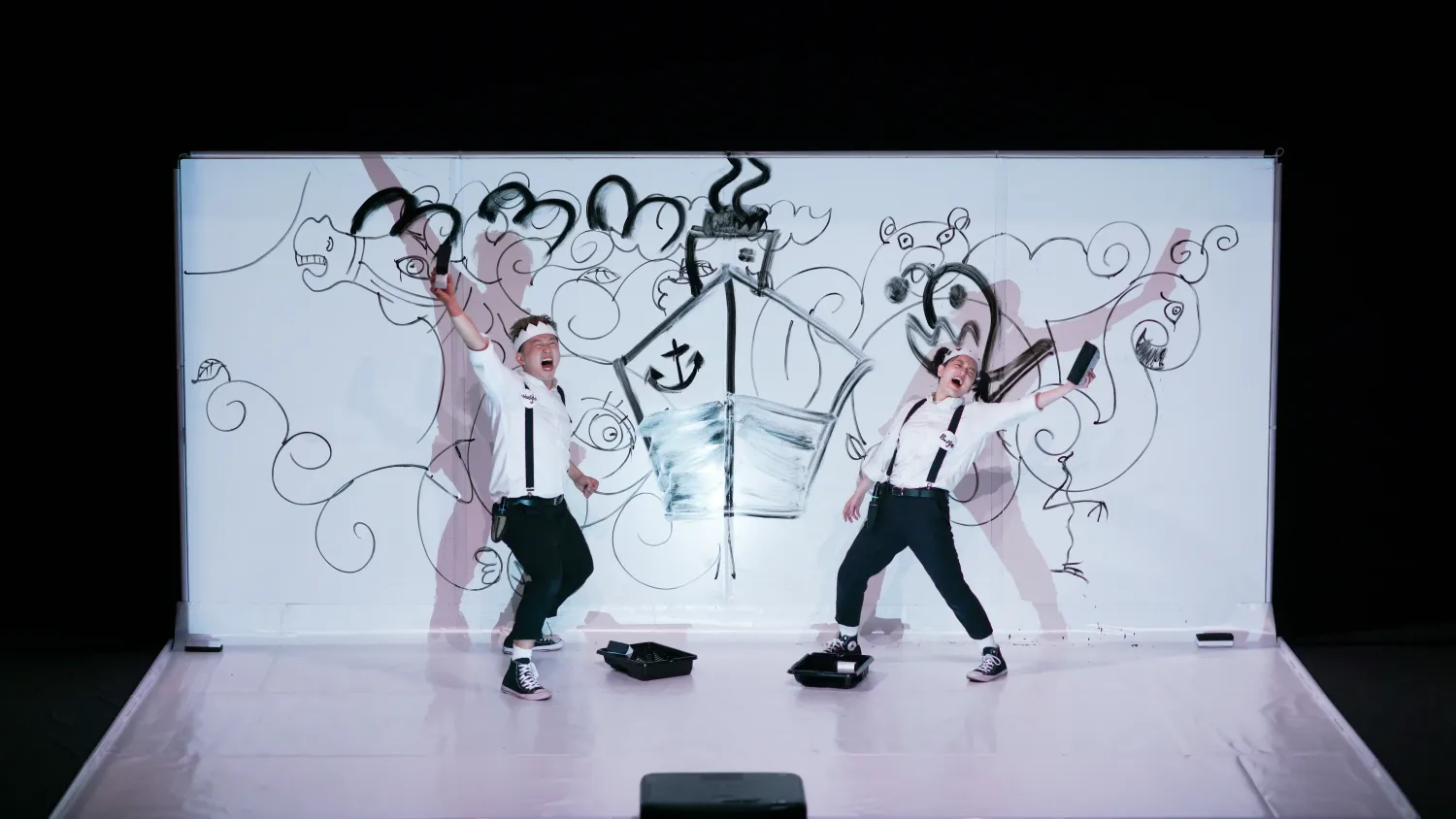 Two performers wearing suspenders and paper crowns exclaim and strike a pose with one arm raised in front of a large white dry erase board.