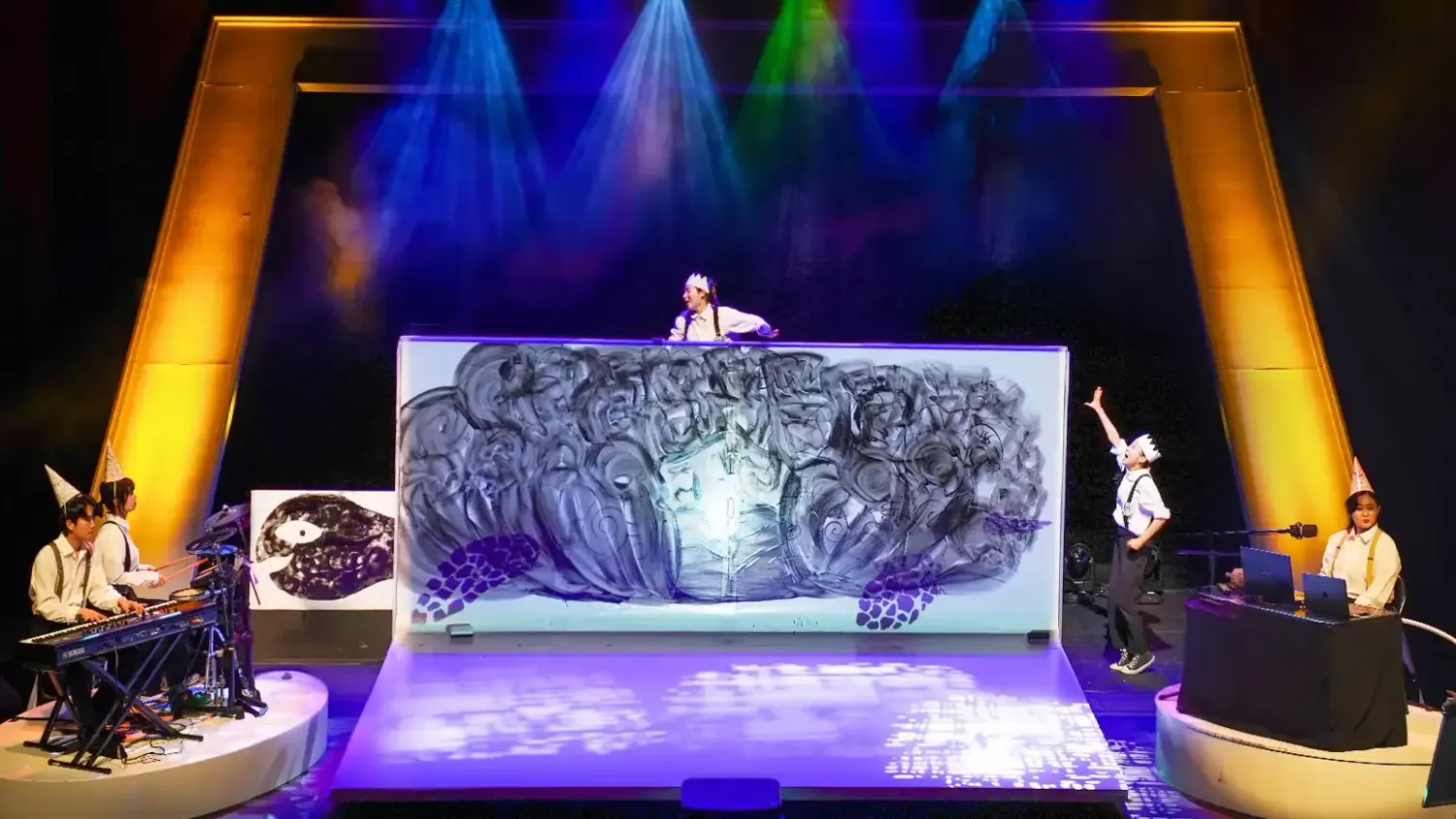 A performer wearing a paper crown rides a large drawing of a turtle. A second performer waves at them. On either side of the stage sit musicians wearing pointed hats.