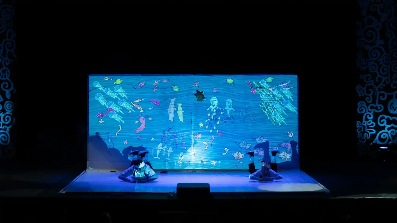 Two performers wearing paper crowns lay on the ground and swim through an ocean full of marine life projected on stage.