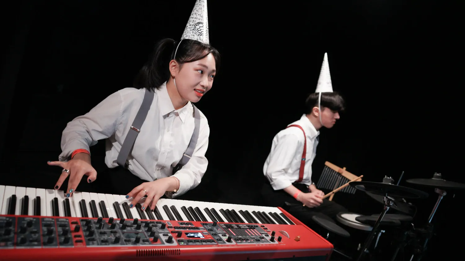 Two musicians wearing suspenders and conical hats play the keyboard and percussion instruments.
