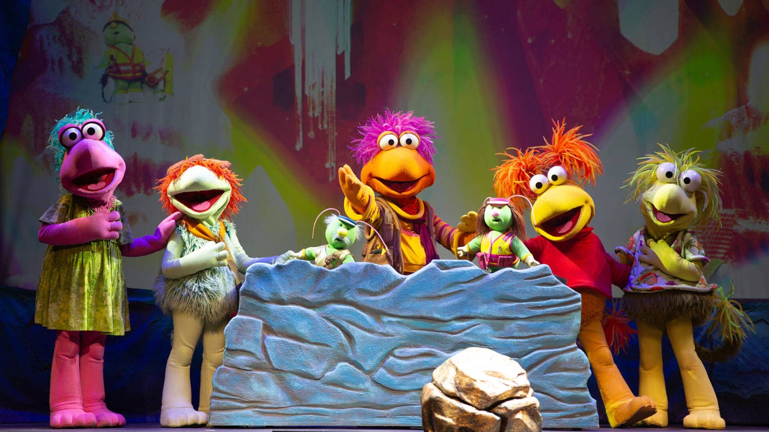 On a stage with a cave backdrop, five colorful Fraggles and two green Doozers stand behind a waist-height wall of stone.