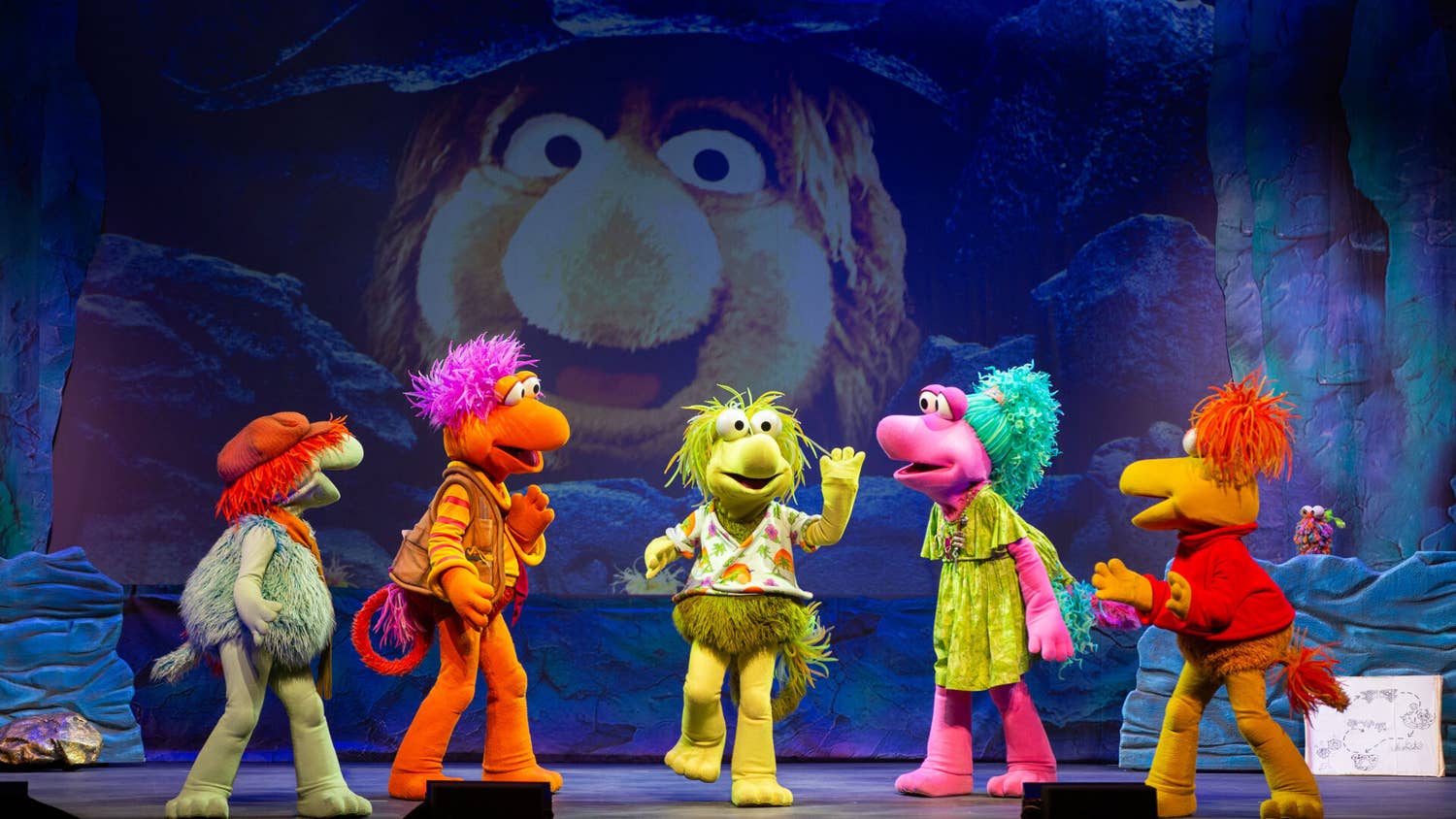 A backdrop shows a large, furry, yellow creature. Five colorful Fraggles stand in a line. The Fraggle in the middle raises one hand and foot.
