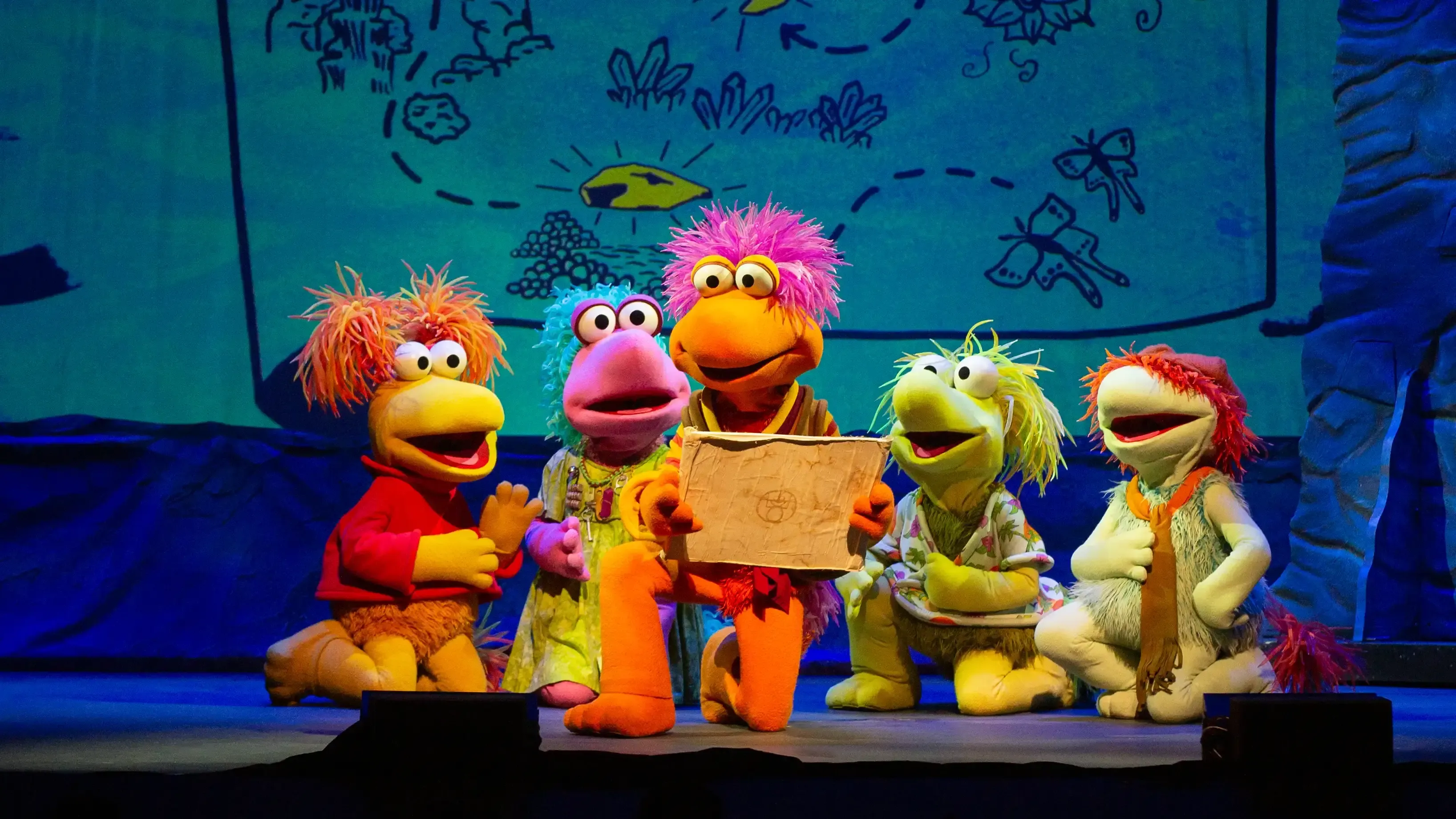 Colorful Fraggle puppet characters with a large box on stage, smiling beneath a graffiti-style backdrop