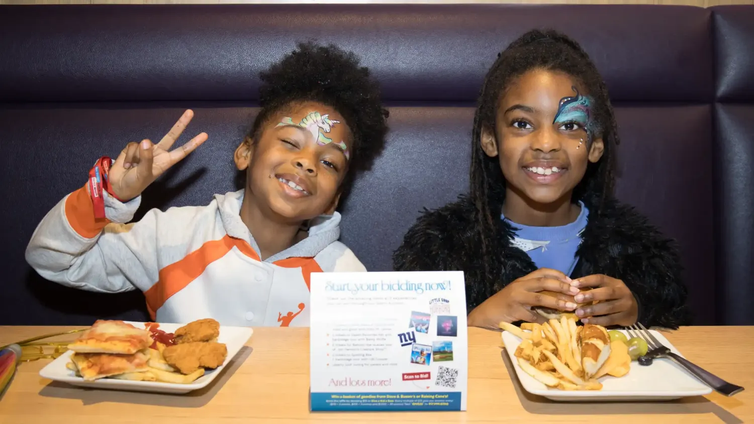 Two kids with face paint smile and eat burgers while sitting in a booth.