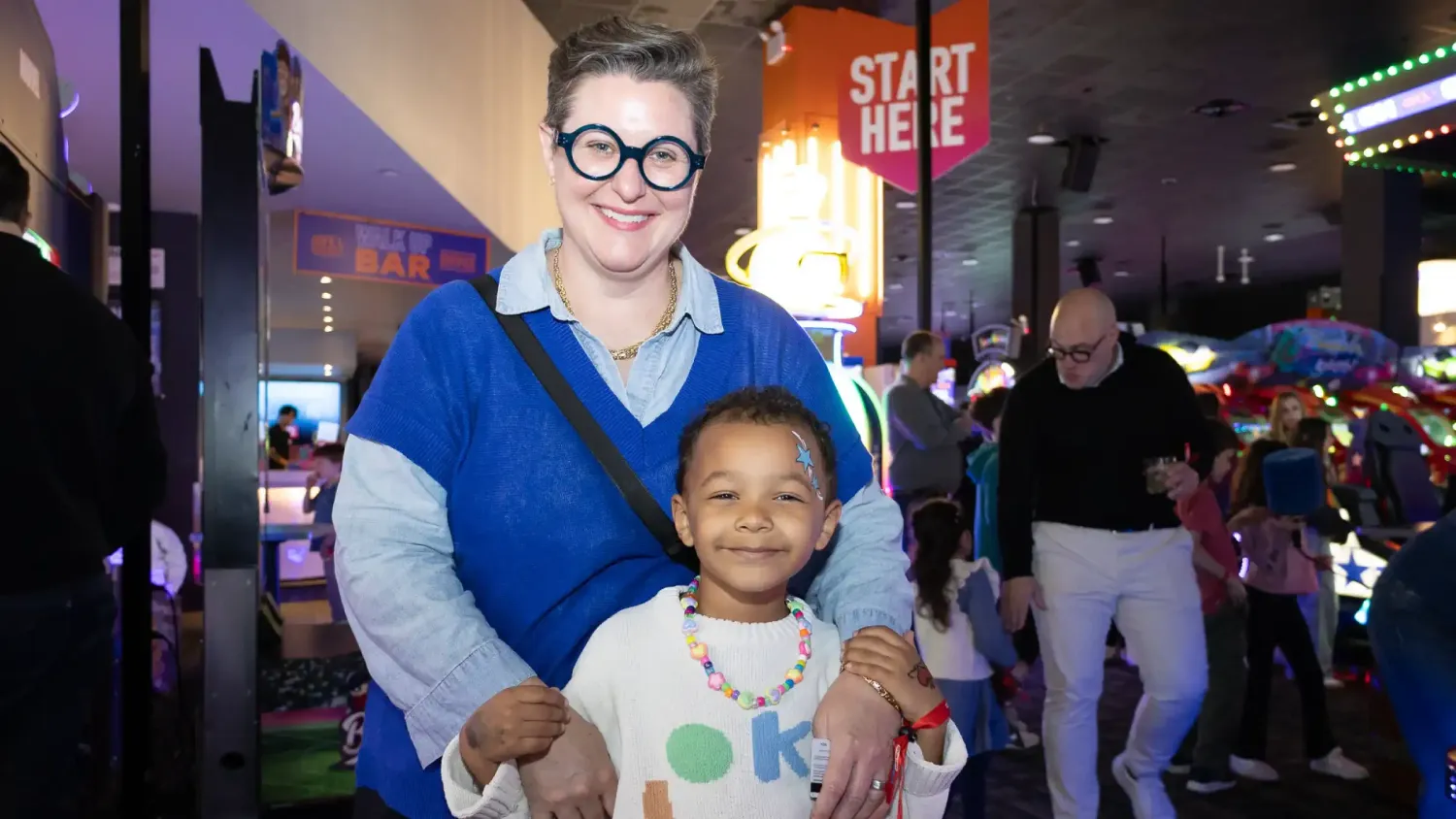 An adult with round glasses and a blue shirt stands behind and holds a kid. They both smile.