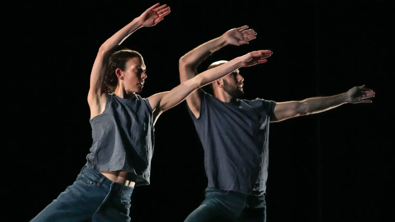 Two dancers in gray shirts and dark jeans face right and hold their arms above their heads, the left arm bent and the right arm straight.