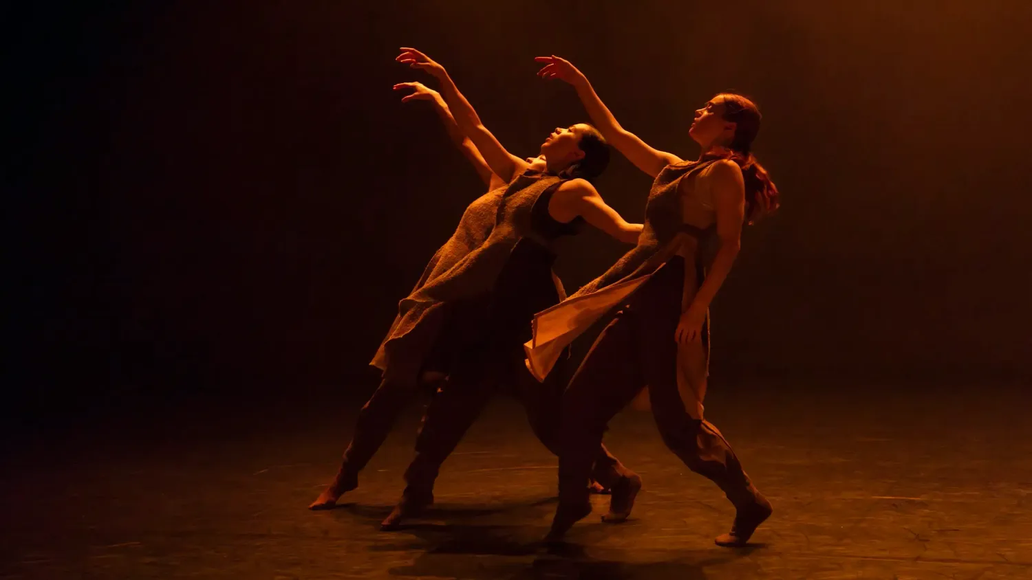 Three dancers lean backwards as they each raise one arm in front of them and look up at their hand. The stage is dimly lit with warm light.
