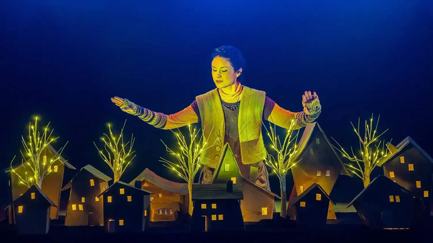 A performer looks down and holds their hands out to their side as they stand over a cardboard village with light-up trees.
