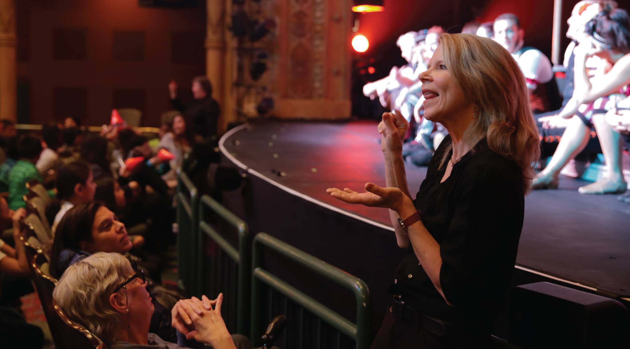 A woman in front of the stage interprets a show with performers out of focus in the background