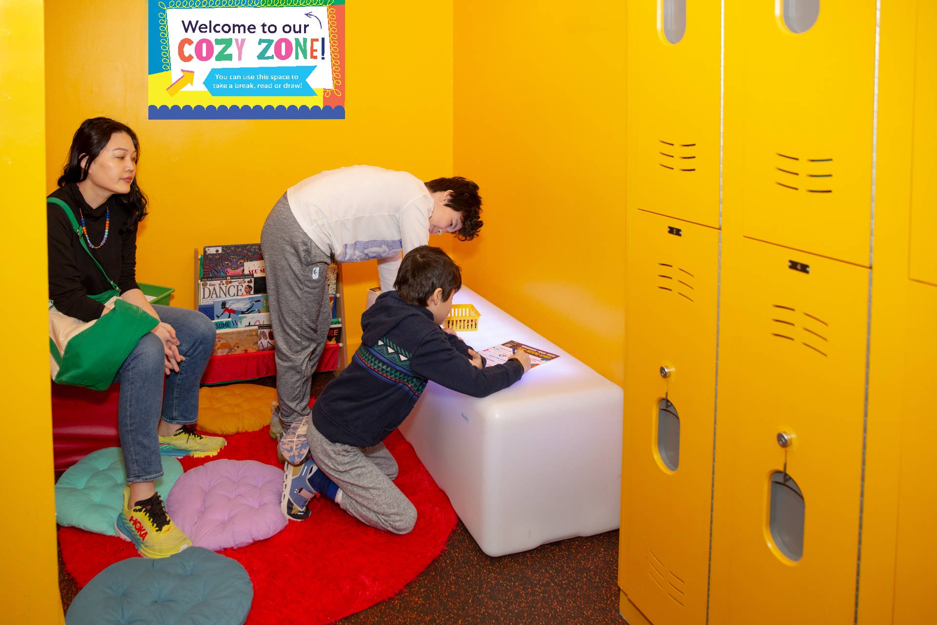 Two kids color and an adult sits and watches in the New Victory Cozy Zone. A sign on the wall behind them reads "Welcome to our Cozy Zone! You can use this space to take a break, read or draw!"