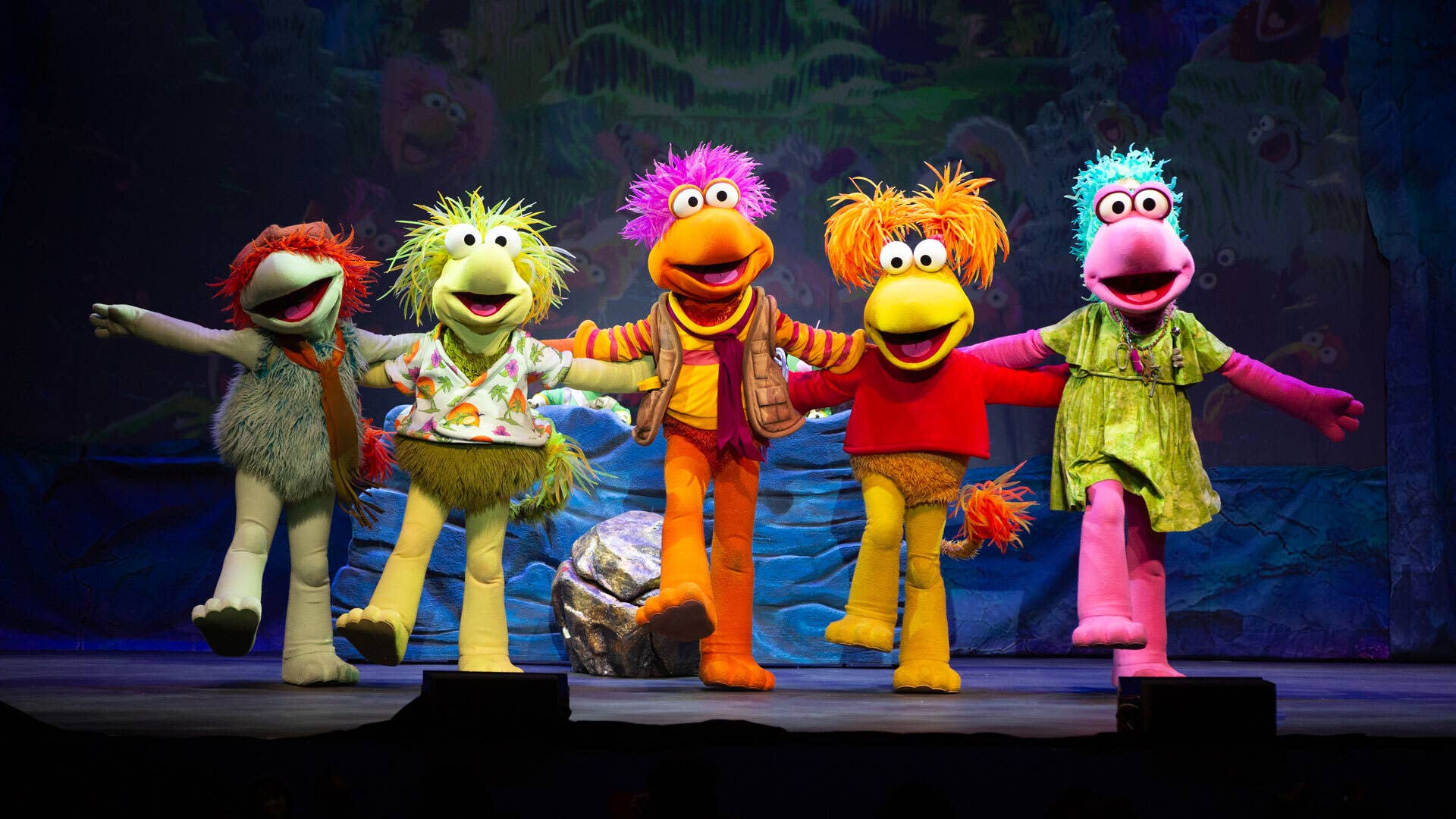 On a stage with a cave backdrop, five Fraggles stand in a kickline with their mouths open, arms outstretched and one foot kicked out in front of them.