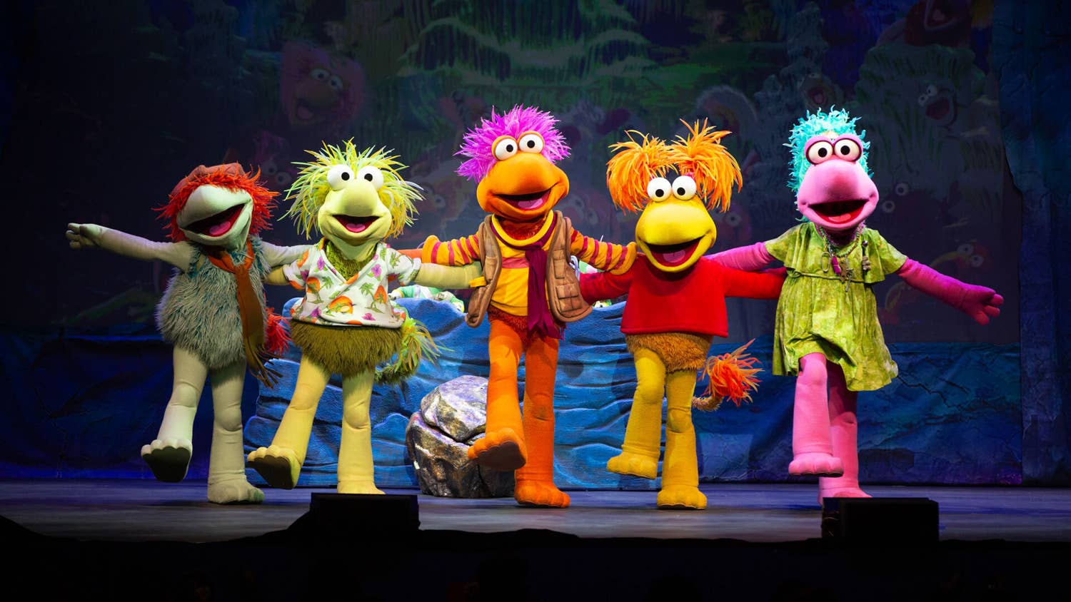 On a stage with a cave backdrop, five Fraggles stand in a kickline with their mouths open, arms outstretched and one foot kicked out in front of them.