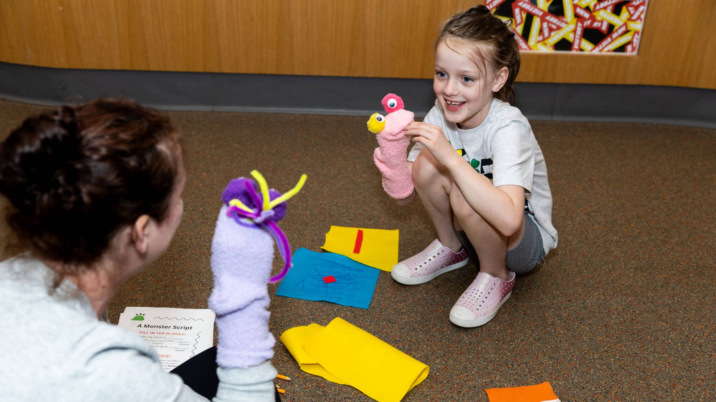 Smiling child kneels in a play area holding a decorated sock during an puppet-making activity