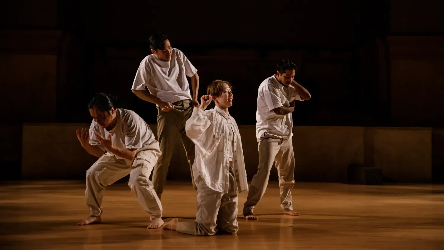 Four dancers in white tops and khakis strike various poses on a stage in low light.