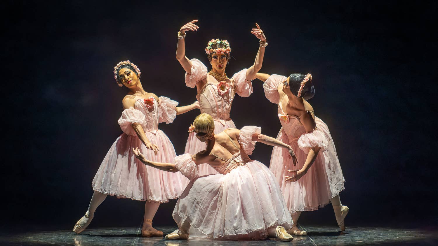 Ballet dancers in pale pink costumes perform a graceful formation with raised arms on a dark stage.