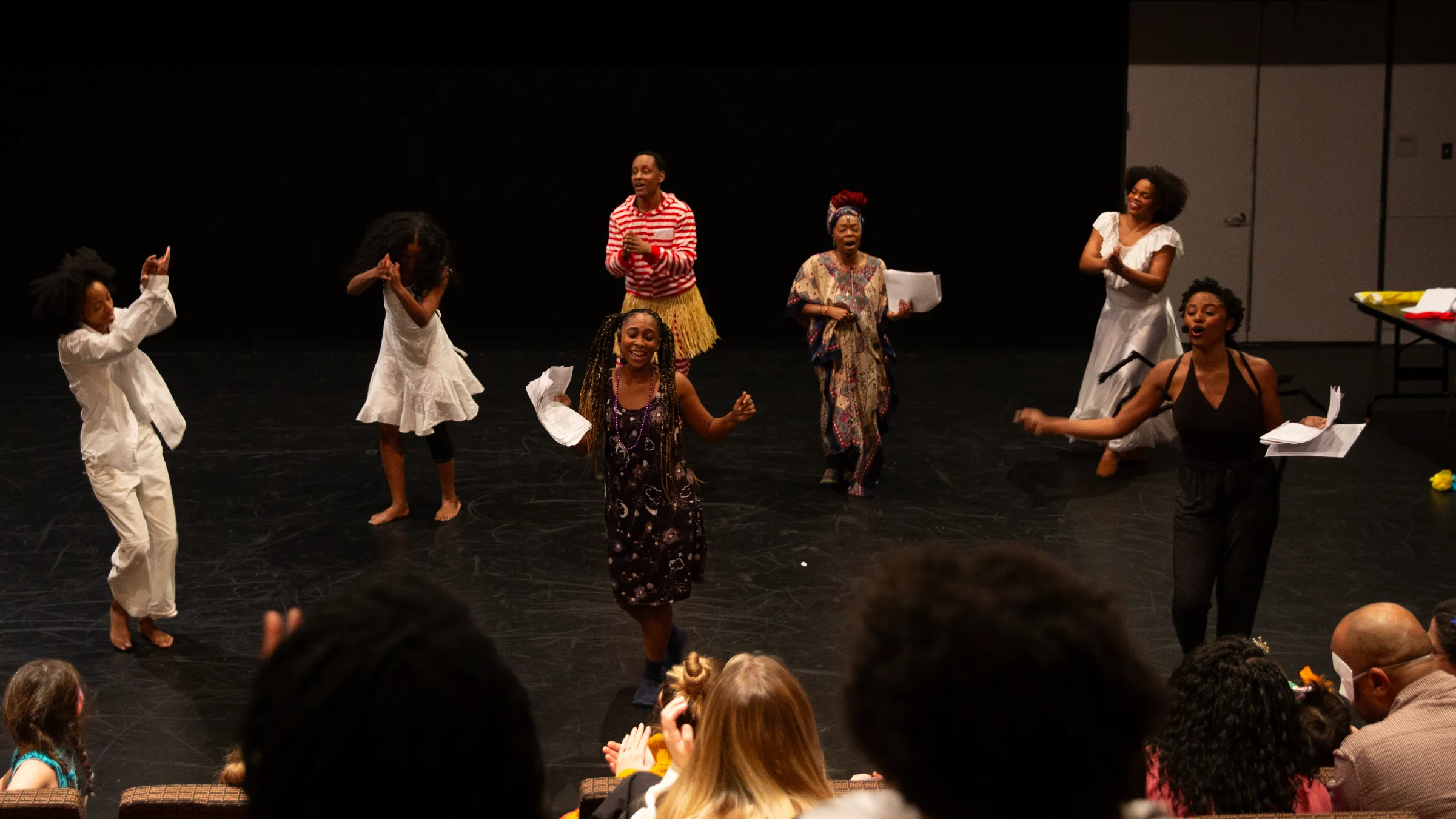 Ensemble of performers on stage in colorful outfits, clapping and singing, with audience in foreground.