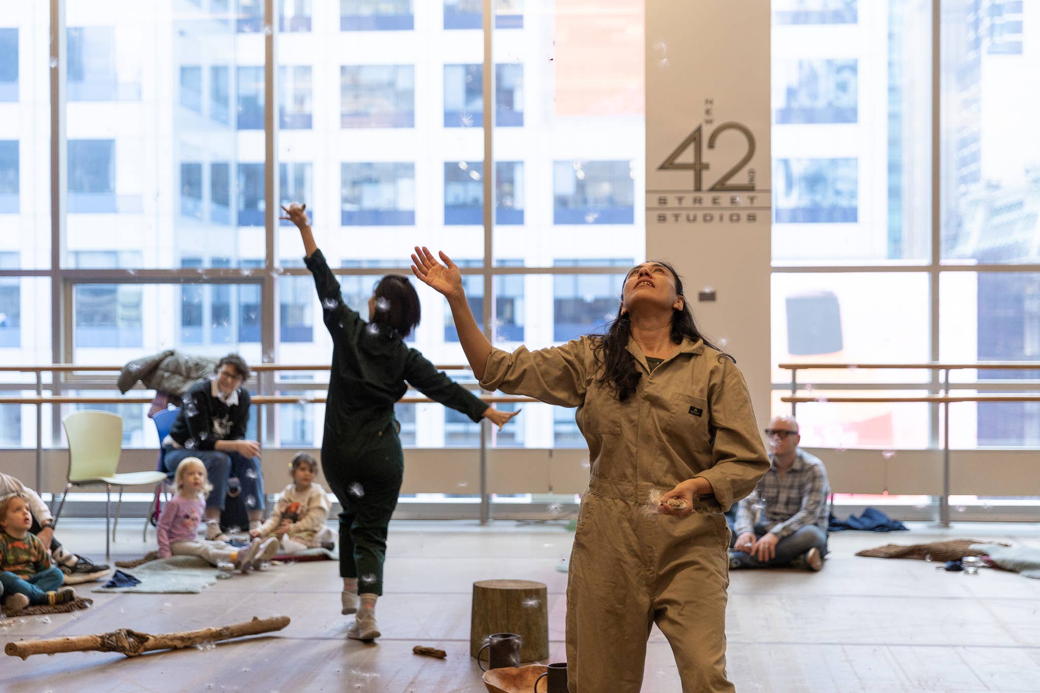 Childcare teacher in tan coveralls leads play, arms up, while kids gather on the floor near windows