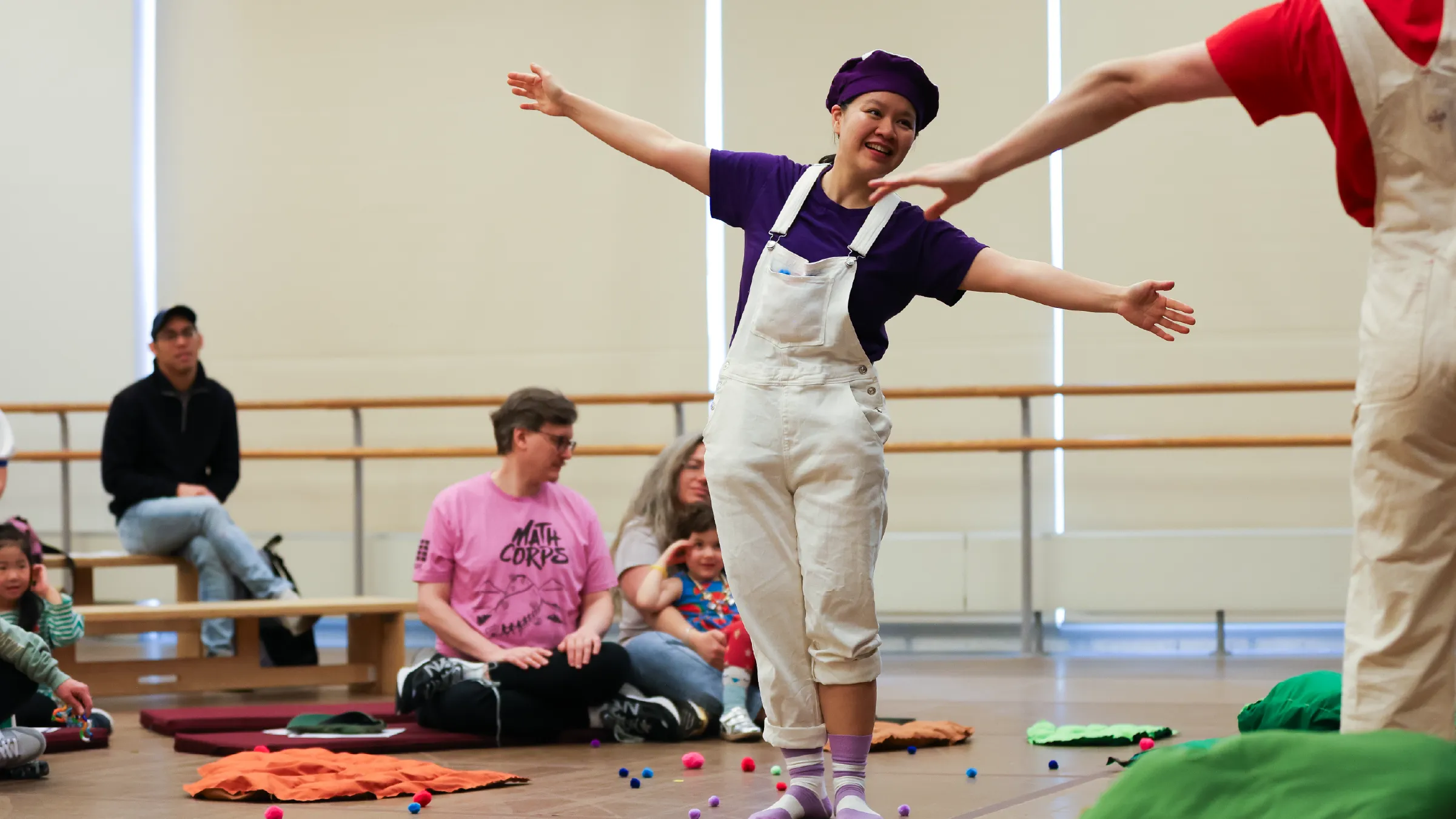 Dancer in purple shirt and white overalls performs with arms out, smiling, in a studio with children watching