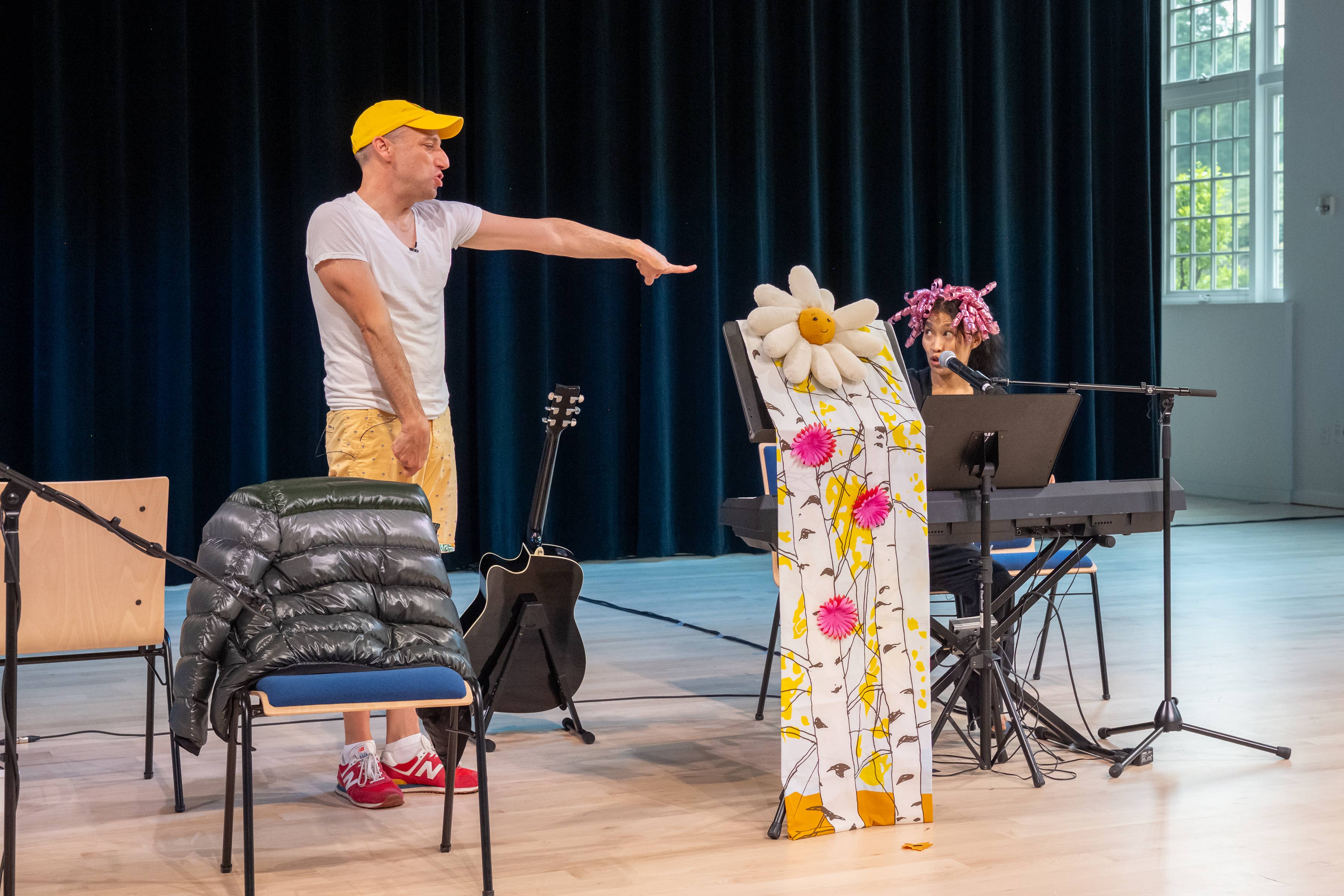 A person in yellow cap and white shirt gestures on stage beside seated performer wearing a floral robe and pink headpiece.
