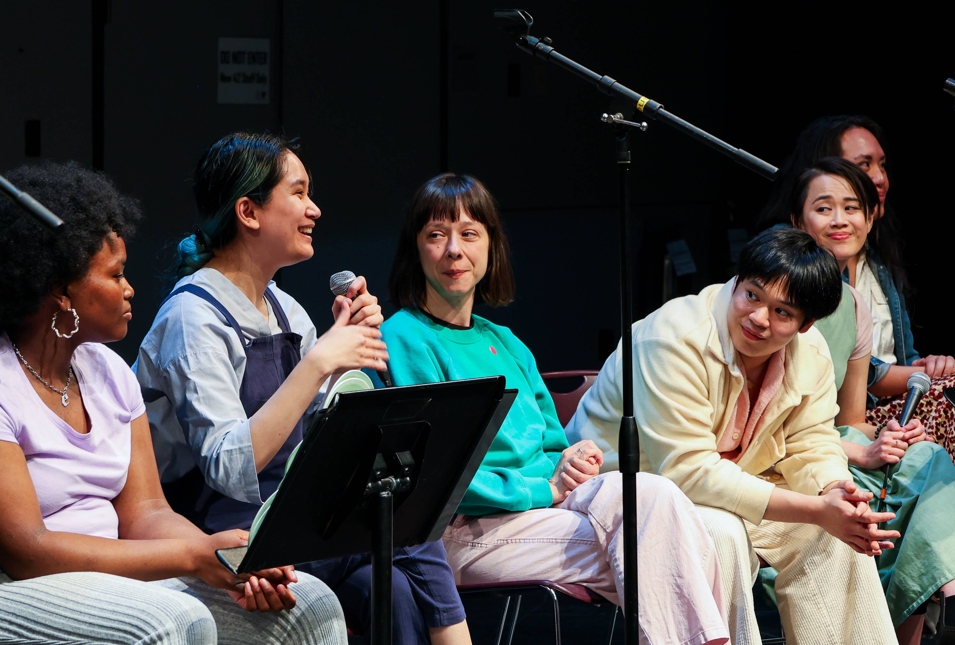 Six people seated in a panel discussion, holding microphones in front of stage lights and a boom mic.