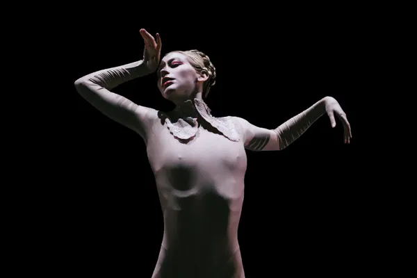 Dancer in a white body suit performs with arms raised against a dark background, head tilted upward.