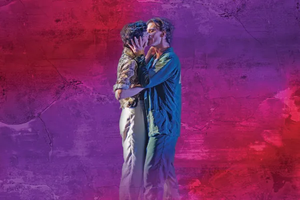 Two men dance and embrace, kissing and smiling against a textured purple and pink wall background.