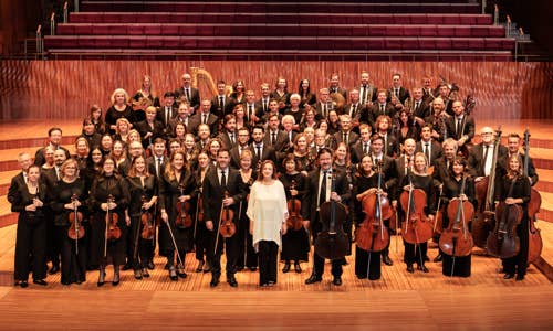 Orchestra group portrait with brass, strings, and woodwinds in a concert hall, posing on stage with instruments.