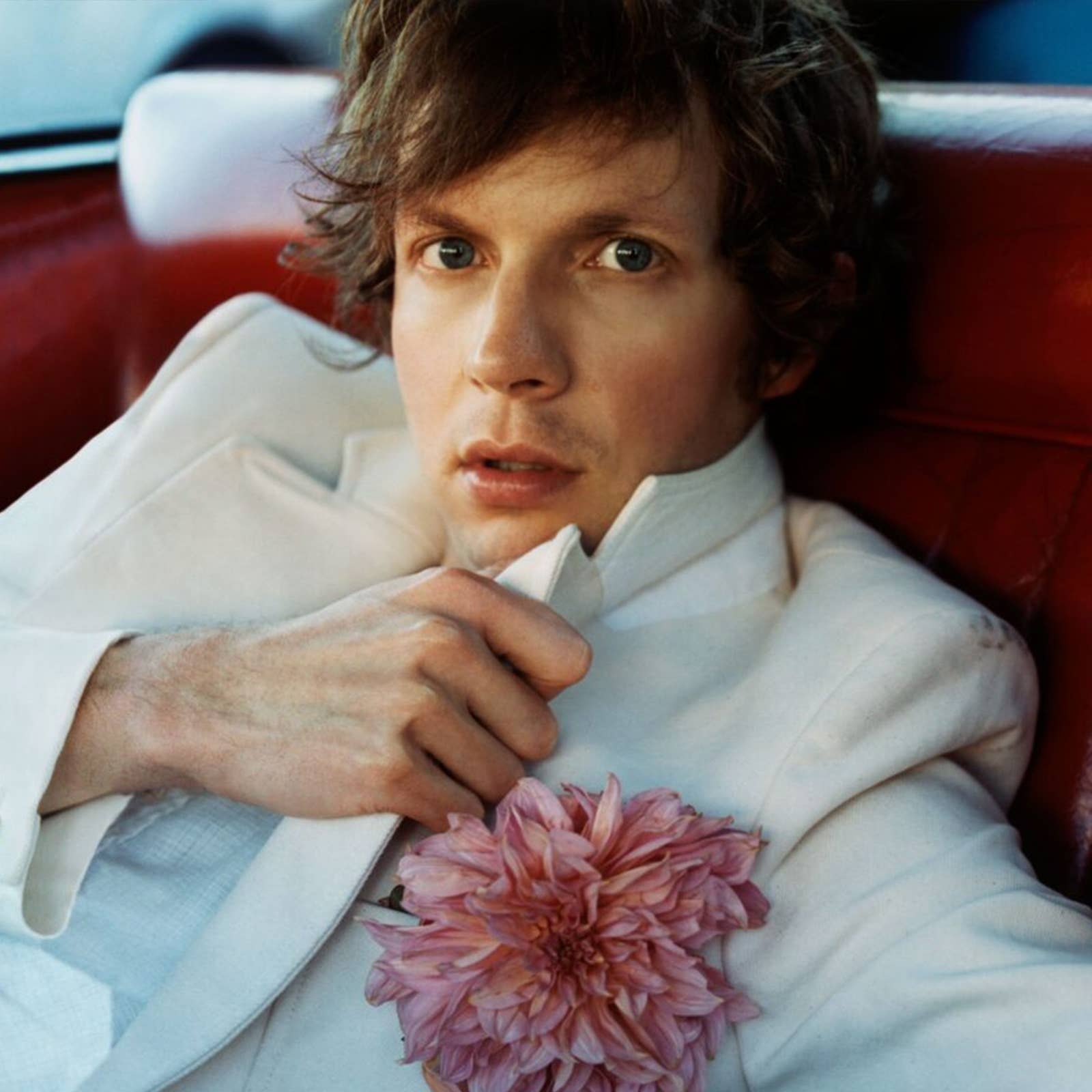 Person in a white suit with a pink flower in the lapel, sitting on a red seat, looking directly at the camera.