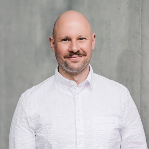 Smiling man with short beard in a white button-up shirt, posed against a light gray wall.