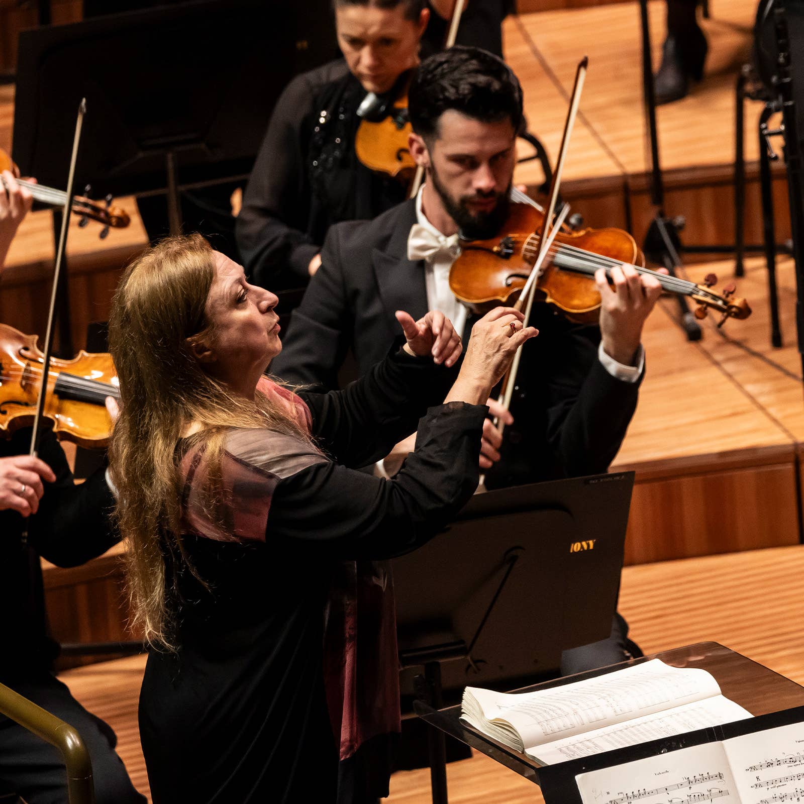 Female conductor and male violinist rehearse with an orchestra in a concert hall under warm lighting.