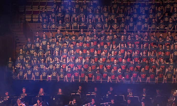 Large choir in matching shirts performs above an orchestra on stage, with a conductor leading the musicians below.