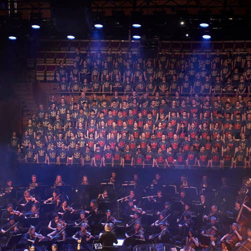 Large choir in matching shirts performs above an orchestra on stage, with a conductor leading the musicians below.